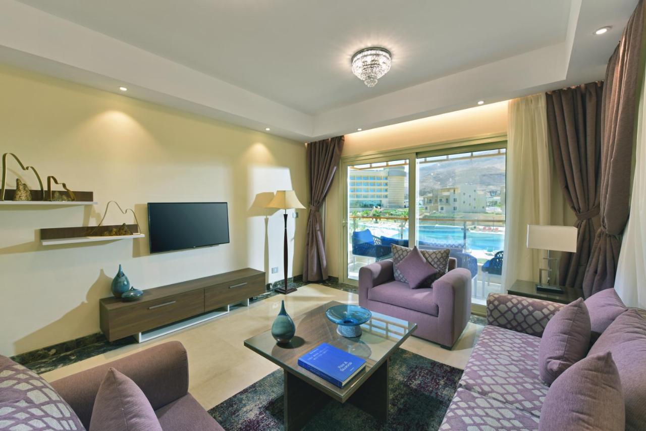 Tolip Resort El Galala Majestic Ain Sukhna Dış mekan fotoğraf The photo shows a modern living room that features a stylish and cozy design. There are two purple sofas with decorative cushions arranged around a coffee table. The coffee table has a blue book and a decorative item on it. A flat-screen TV is mounte