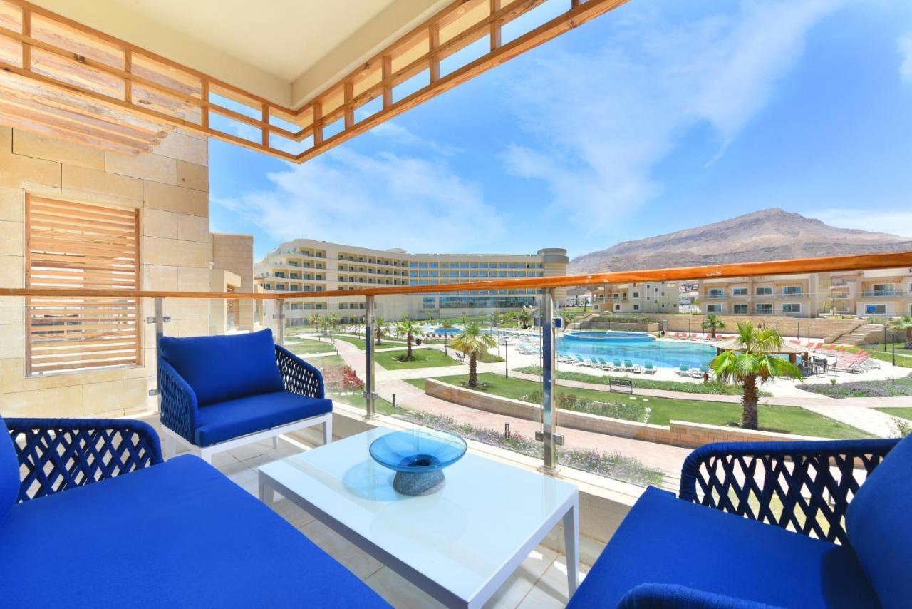 Tolip Resort El Galala Majestic Ain Sukhna Dış mekan fotoğraf The photo showcases a balcony view featuring two blue chairs and a table with a decorative bowl. The balcony overlooks a landscape that includes a swimming pool and manicured lawns. In the background, there are multiple buildings and palm trees, all 