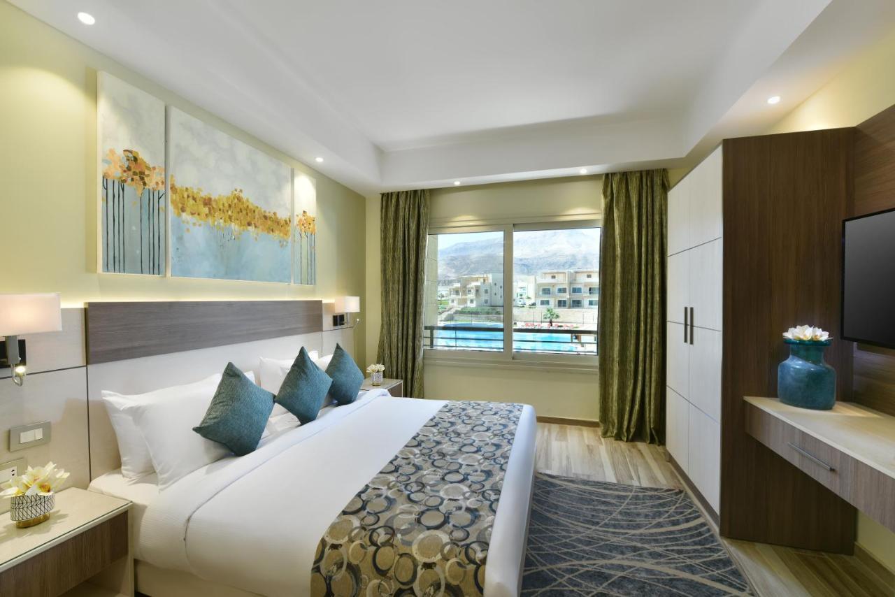 Tolip Resort El Galala Majestic Ain Sukhna Dış mekan fotoğraf The photo shows a modern hotel room. It features a large bed with white bedding and several decorative pillows in shades of teal. Above the bed, there are two pieces of wall art with a landscape theme. The room has a large window with green curtains,