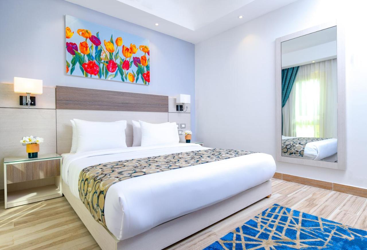 Tolip Resort El Galala Majestic Ain Sukhna Dış mekan fotoğraf The photo shows a modern hotel bedroom. It features a large bed with white linens and a decorative throw blanket. The room has light wooden flooring and is bright, with ample natural light coming from a window. There is a colorful painting of flowers
