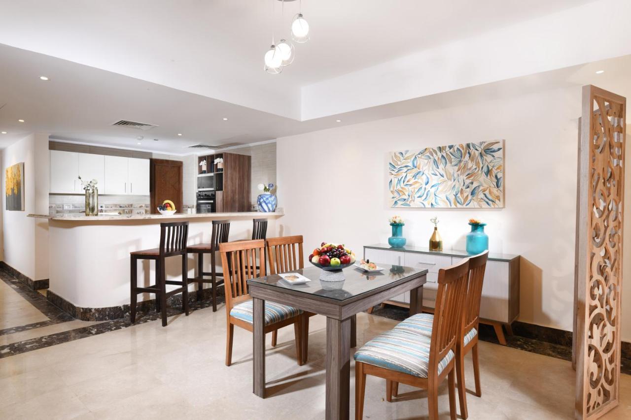 Tolip Resort El Galala Majestic Ain Sukhna Dış mekan fotoğraf The photo shows a modern dining area with a small table and chairs. There is a centerpiece with a bowl of fruit on the table. The room features a light-colored floor and walls, and there is artwork with a colorful design on one wall. In the backgroun