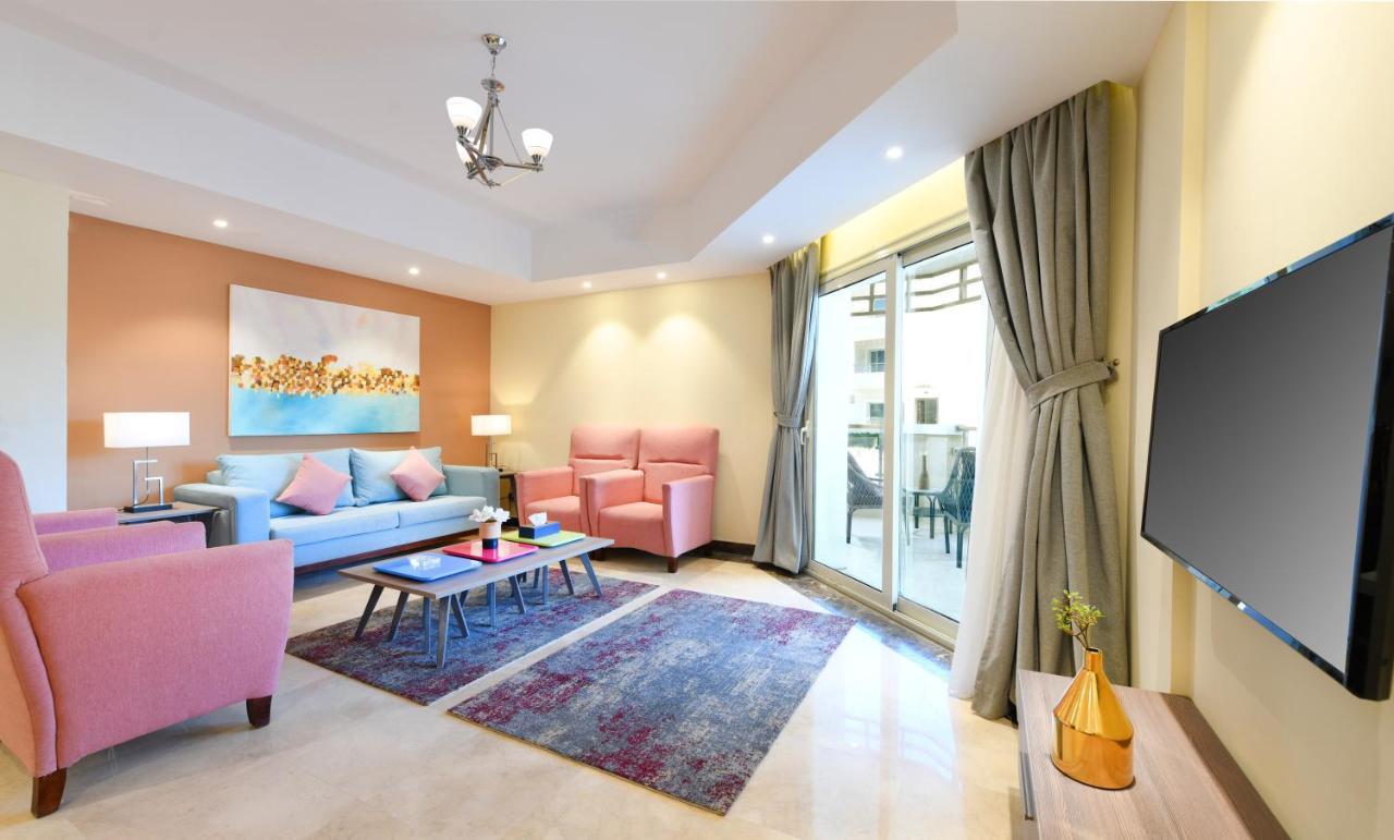 Tolip Resort El Galala Majestic Ain Sukhna Oda fotoğraf The photo shows a modern living room that features a cozy and stylish design. There is a light blue sofa adorned with pastel pink cushions, accompanied by two pink armchairs. A coffee table sits in front, placed on a colorful area rug that adds warmt