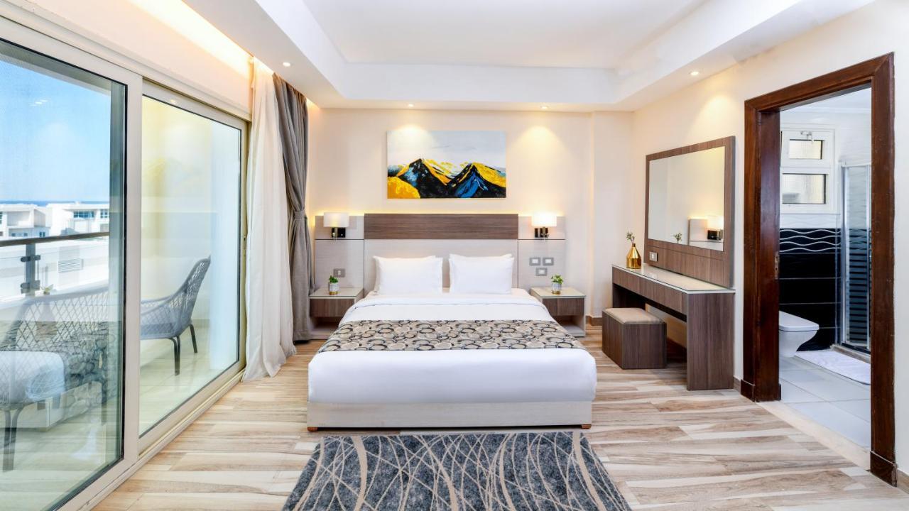 Tolip Resort El Galala Majestic Ain Sukhna Dış mekan fotoğraf The photo shows a modern hotel room featuring a large bed with white linens and a decorative throw. The room has a neutral color palette, accented with warm lighting. There is a piece of artwork above the bed depicting mountains. To the left, there a