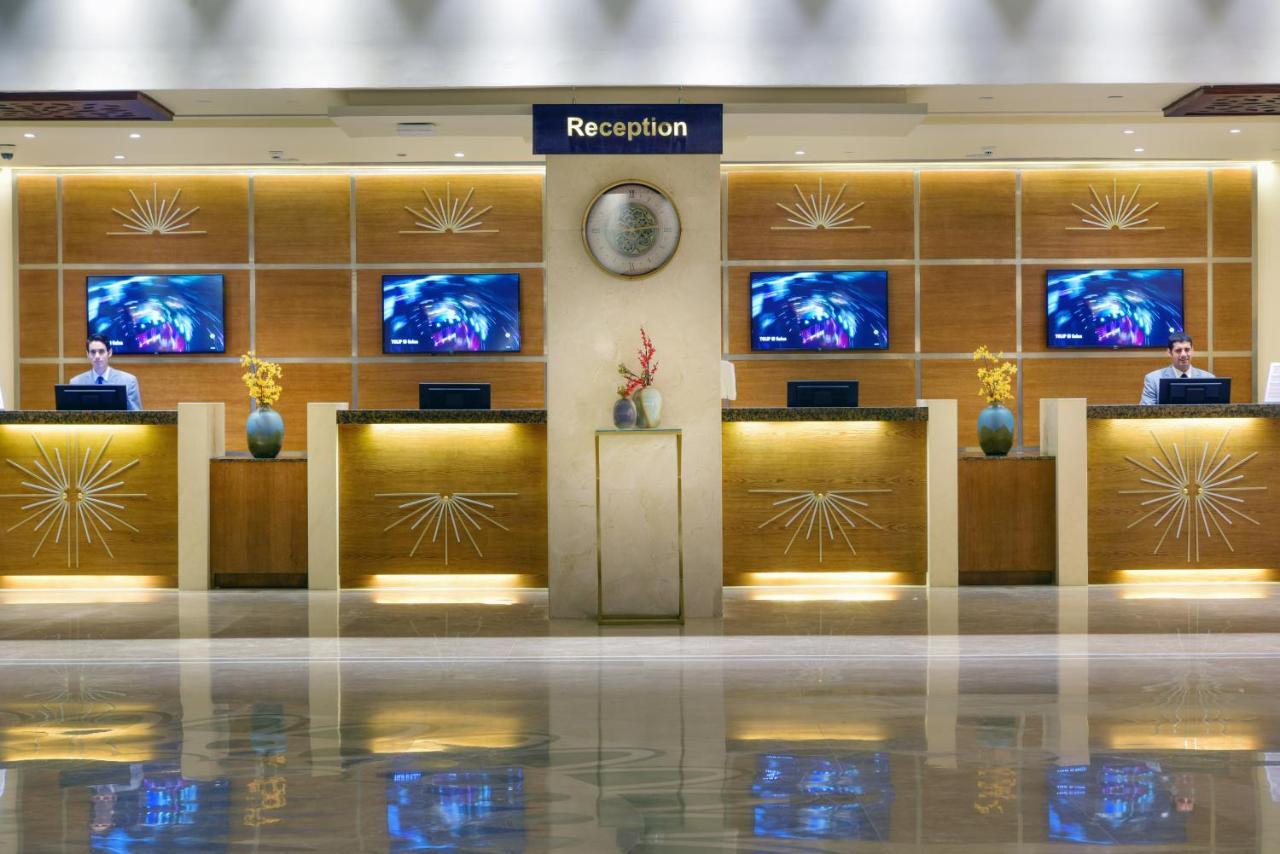 Tolip Resort El Galala Majestic Ain Sukhna Dış mekan fotoğraf The image shows a hotel reception area. There are several reception desks lined up against a wall with decorative paneling. Each desk has a staff member, and above the desks, there are screens displaying information. A clock is visible on the wall, a