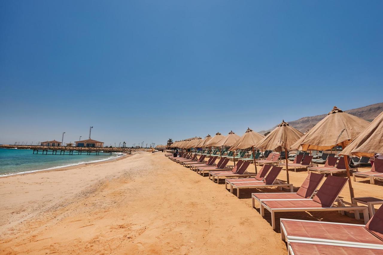 Tolip Resort El Galala Majestic Ain Sukhna Dış mekan fotoğraf The photo shows a scenic beach with a stretch of golden sand under a clear blue sky. There are several beach loungers arranged in rows, each accompanied by thatched umbrellas providing shade. In the background, there is a wooden pier extending into t