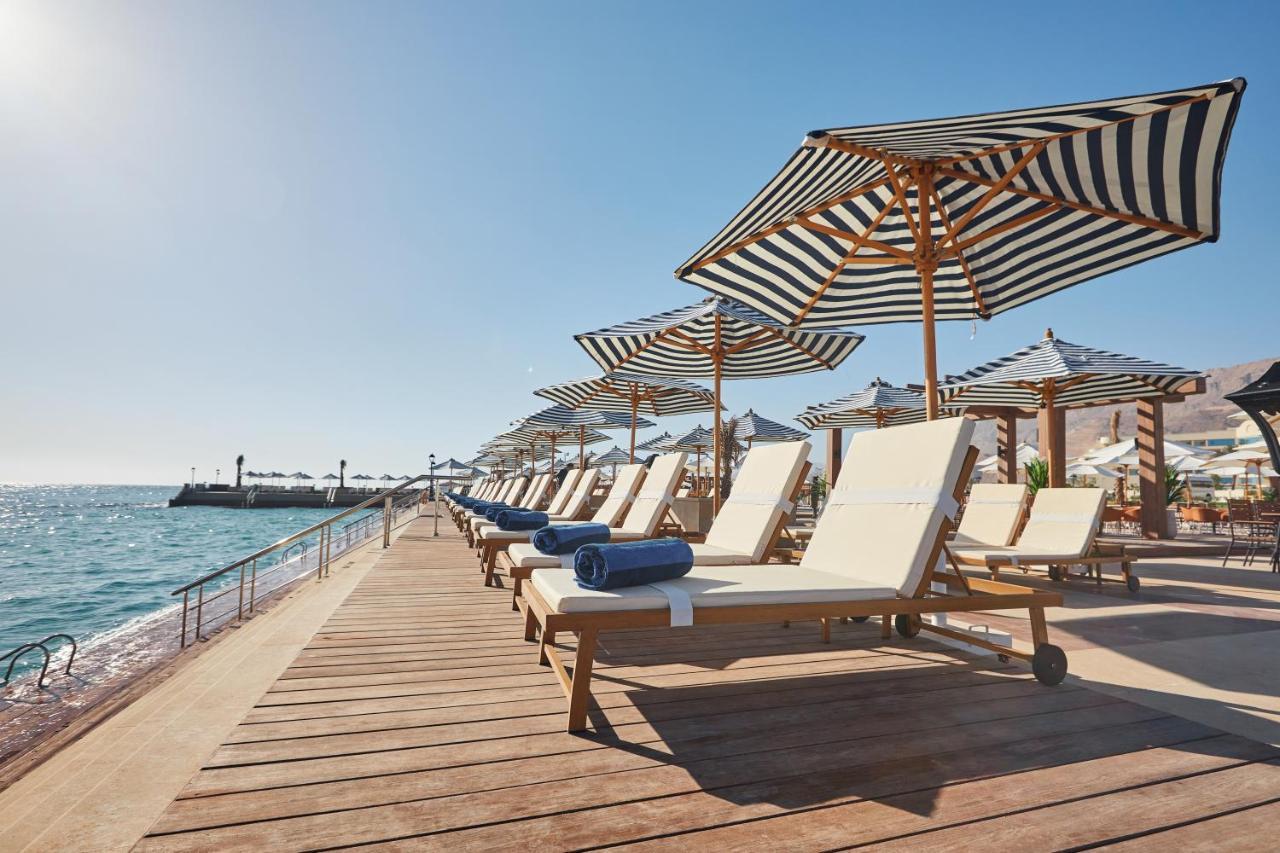 Tolip Resort El Galala Majestic Ain Sukhna Dış mekan fotoğraf The photo depicts a serene beachside setting featuring a row of sun loungers arranged on a wooden deck. Each lounger is equipped with a folded towel and shaded by striped umbrellas. In the background, the water sparkles under bright sunlight, and the