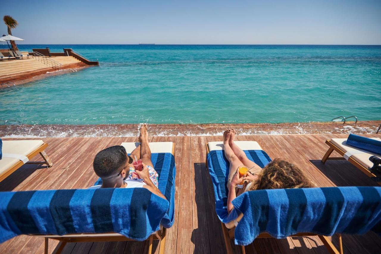 Tolip Resort El Galala Majestic Ain Sukhna Dış mekan fotoğraf The photo depicts two people relaxing on lounge chairs by the waterfront. They are positioned on a wooden deck with a view of the clear, turquoise water. Both individuals are reclining, enjoying the sun, and each has a drink in hand. The scene convey