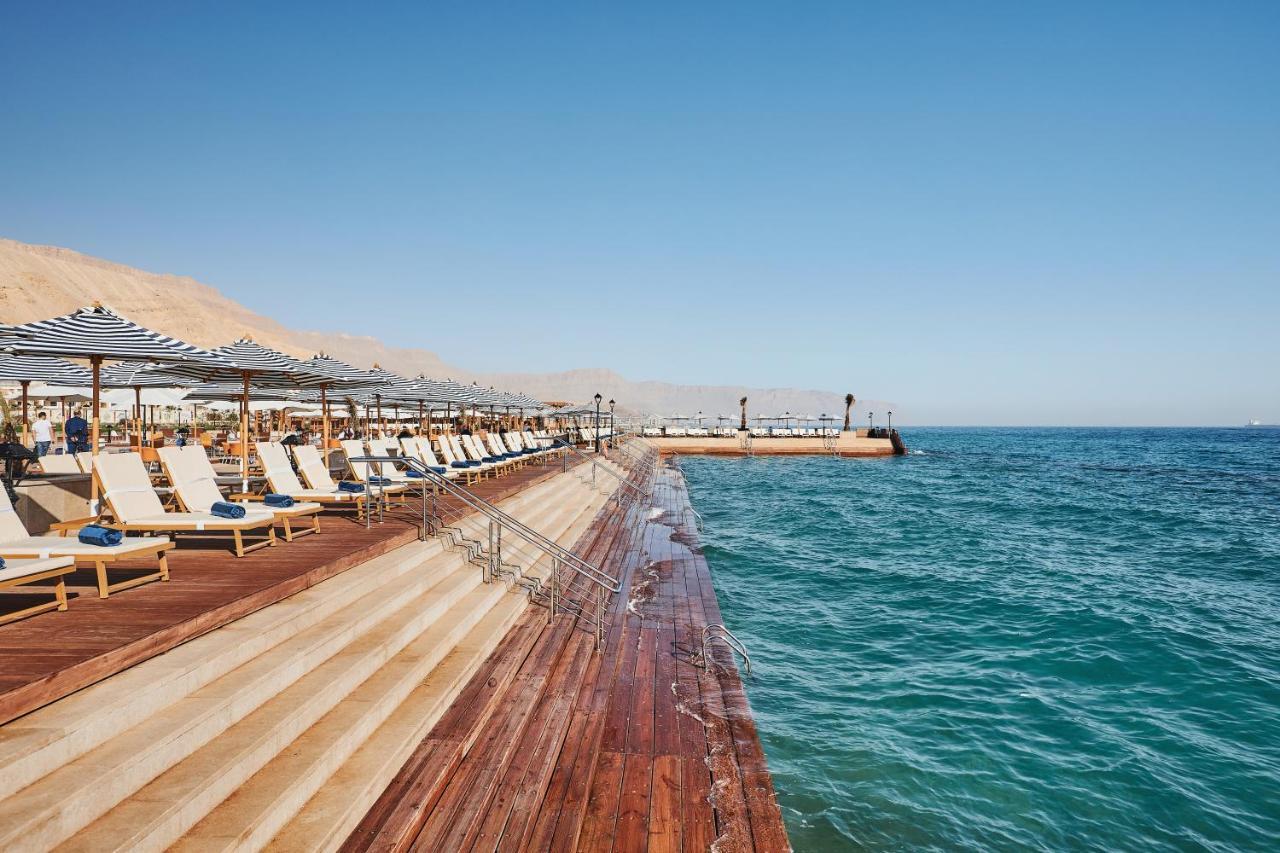 Tolip Resort El Galala Majestic Ain Sukhna Dış mekan fotoğraf The photo shows a seaside area featuring a wooden deck that extends into the water. On the left side, there are several lounge chairs arranged neatly under shade structures, suggesting a relaxing atmosphere. The ocean is visible in the foreground, wi