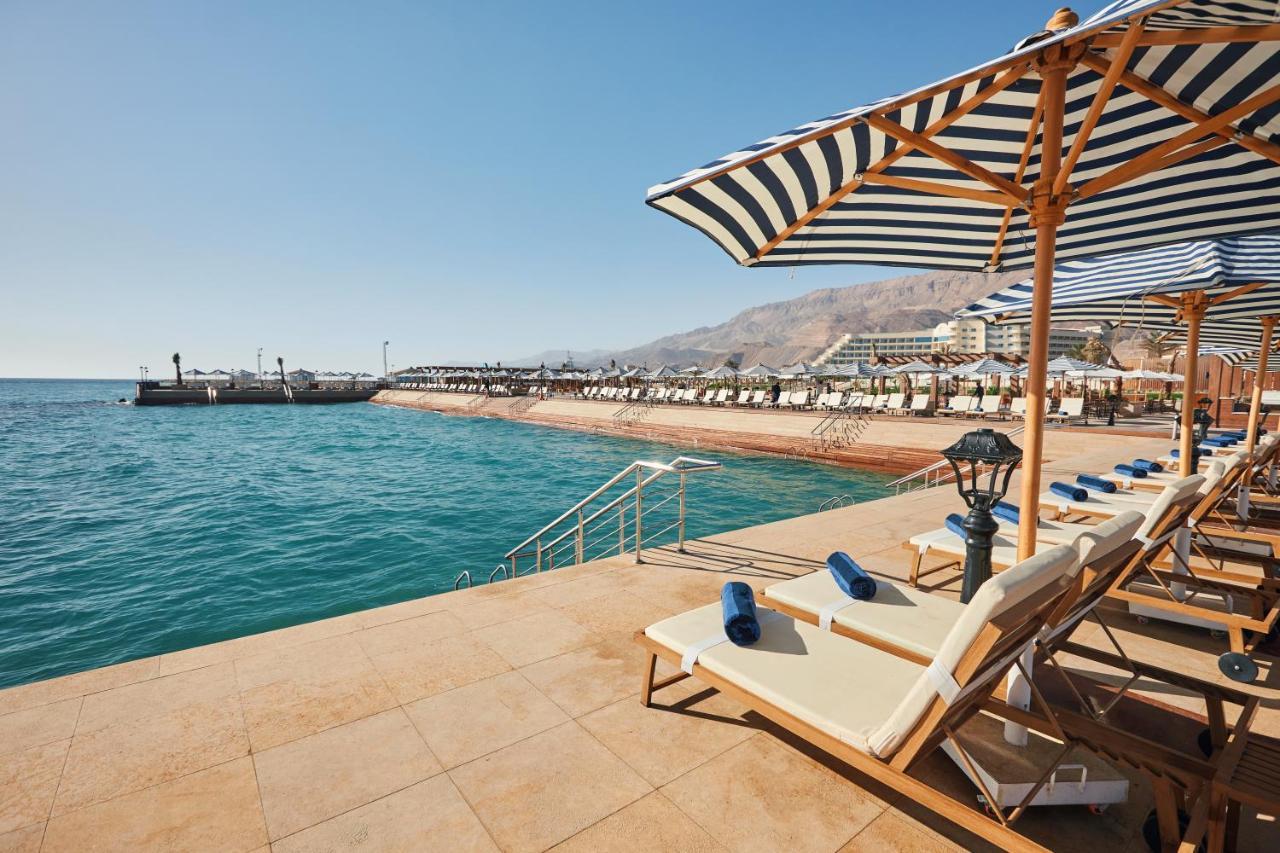 Tolip Resort El Galala Majestic Ain Sukhna Dış mekan fotoğraf The photo shows a scenic beachside area with clear blue water. In the foreground, there are several sun loungers set up on a tiled surface, with blue towels neatly rolled on each one. The loungers are shaded by striped umbrellas. In the distance, a w