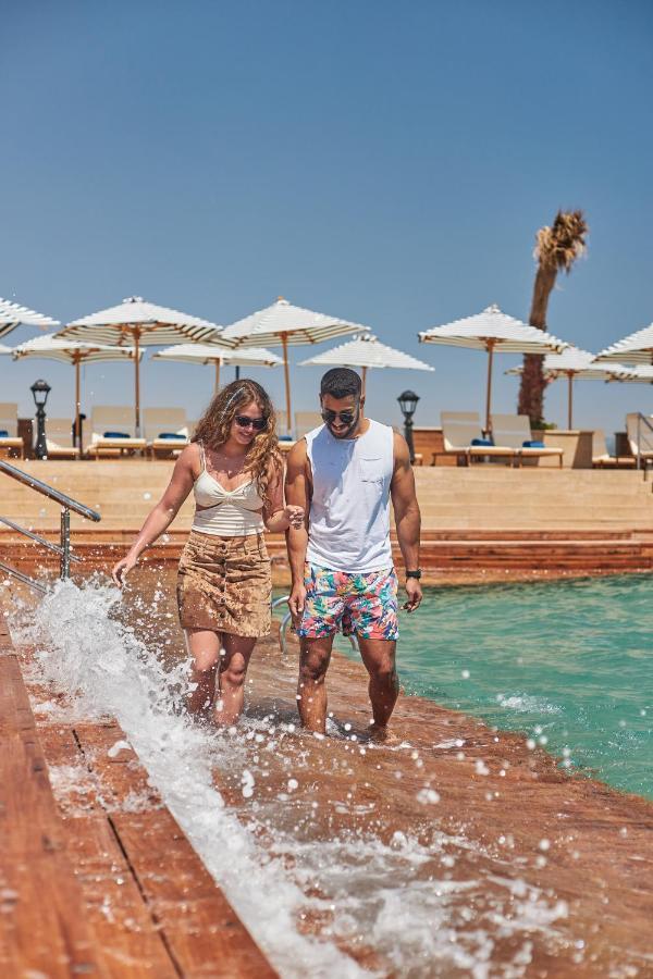 Tolip Resort El Galala Majestic Ain Sukhna Dış mekan fotoğraf The photo depicts a couple walking along a poolside area. They are enjoying a sunny day near a swimming pool, with splashes of water around them. The woman is wearing a light-colored top and a patterned skirt, while the man is dressed in a sleeveless