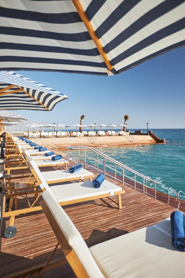 Tolip Resort El Galala Majestic Ain Sukhna Dış mekan fotoğraf The photo depicts a serene outdoor area by the waterfront. There are several lounge chairs arranged in a row, each equipped with blue towels. Large, striped umbrellas provide shade over some of the chairs. In the background, there is a wooden deck th