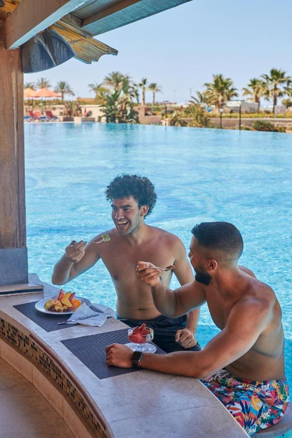Tolip Resort El Galala Majestic Ain Sukhna Dış mekan fotoğraf The photo features two young men enjoying a meal together. They are sitting at a poolside table with a clear blue swimming pool in the background. The men are shirtless, suggesting a casual, relaxed atmosphere, likely in a warm environment. One man i
