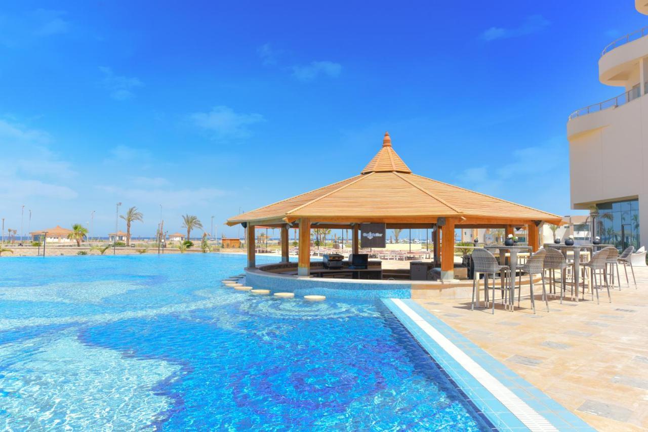 Tolip Resort El Galala Majestic Ain Sukhna Dış mekan fotoğraf The photo shows a beautiful outdoor setting featuring a large swimming pool with clear blue water. In the center, there is a thatched-roof gazebo, which serves as a shaded seating area. Surrounding the gazebo are lounge chairs and a bar area with hig