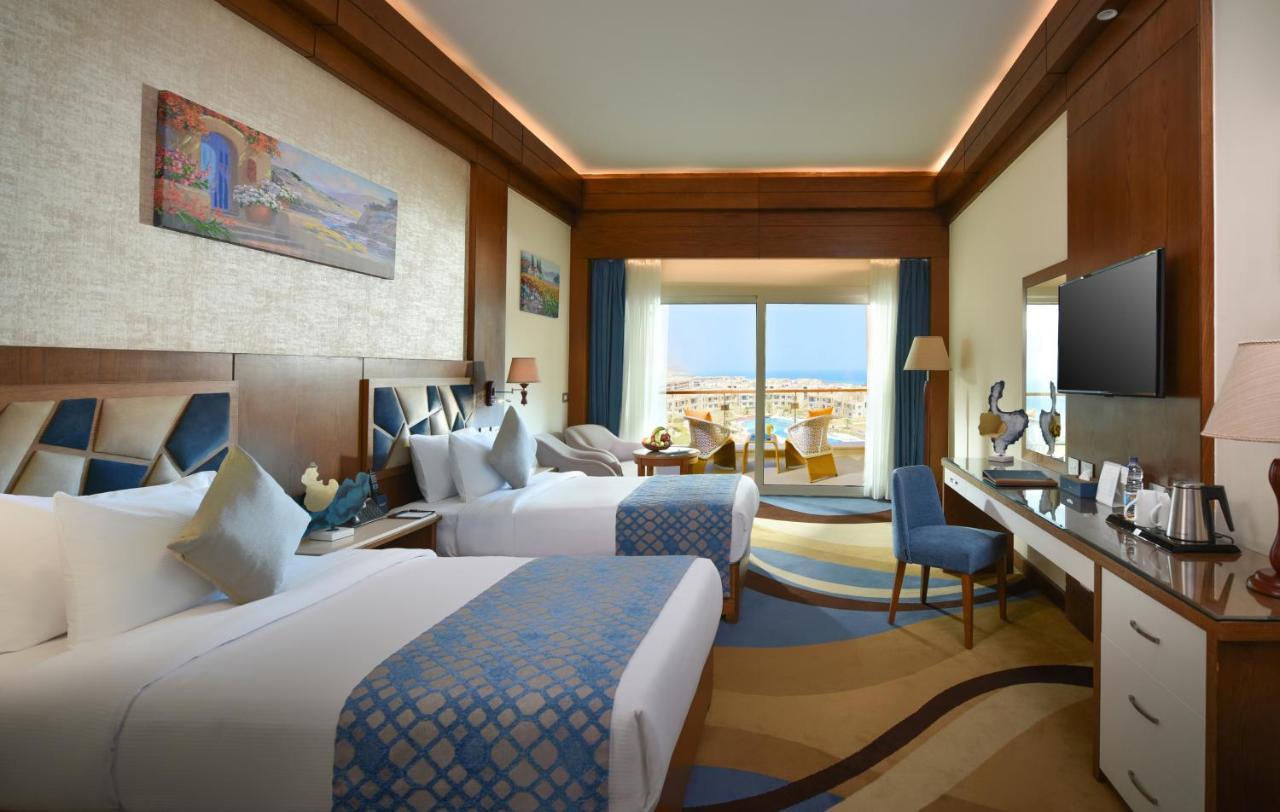 Tolip Resort El Galala Majestic Ain Sukhna Dış mekan fotoğraf The photo shows a well-decorated hotel room featuring two beds with decorative pillows. The room has wooden accents and modern furnishings, including a desk, a television, and lamps. Large windows flood the space with natural light, offering a view o