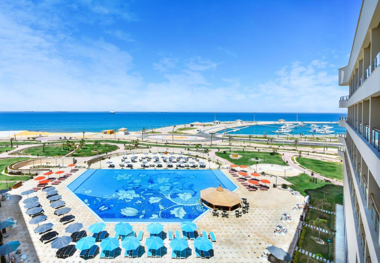 Tolip Resort El Galala Majestic Ain Sukhna Dış mekan fotoğraf The photo shows a beautiful outdoor view of a resort area featuring a large swimming pool with a decorative underwater design. Surrounding the pool are numerous sun loungers and umbrellas in a variety of colors. In the background, there is a scenic c