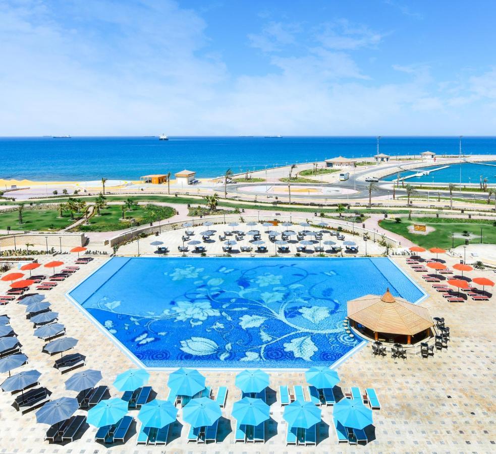 Tolip Resort El Galala Majestic Ain Sukhna Dış mekan fotoğraf The photo features a large swimming pool with a decorative design on its surface, surrounded by sun loungers and umbrellas. In the background, there is a view of the sea, with a clear blue sky and a few scattered clouds. The area is well-maintained, 