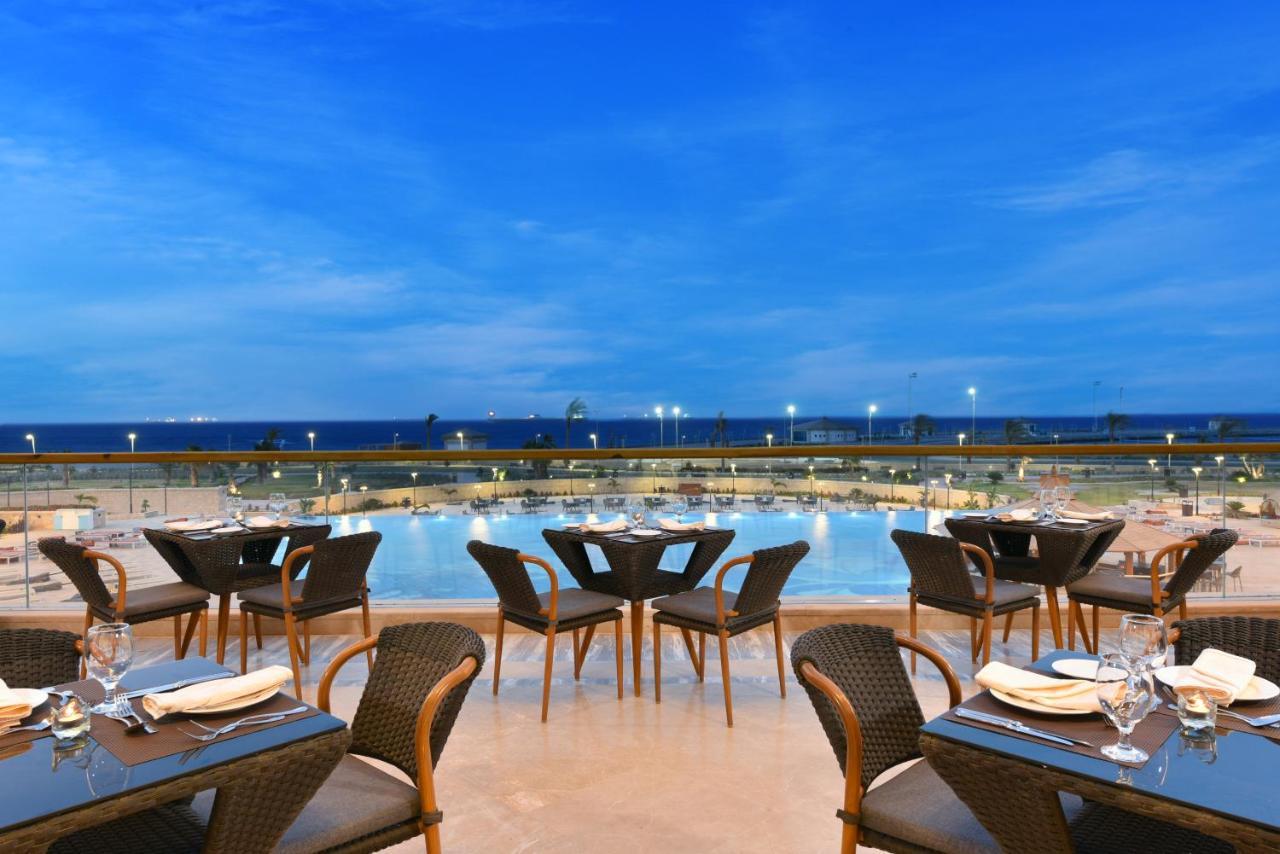 Tolip Resort El Galala Majestic Ain Sukhna Dış mekan fotoğraf The photo shows an outdoor dining area with several neatly arranged tables and chairs. The tables are elegantly set with cutlery and plates, suggesting a fine dining experience. In the background, there is a large swimming pool that reflects the blue