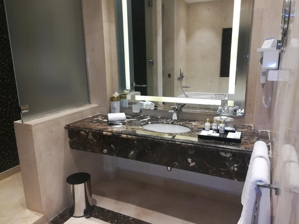 Tolip Resort El Galala Majestic Ain Sukhna Dış mekan fotoğraf The photo shows a modern bathroom with elegant design features. There is a large vanity made of dark marble with a sink and a set of toiletries neatly arranged on top. A mirror with built-in lighting is mounted above the sink. To the side, there is a