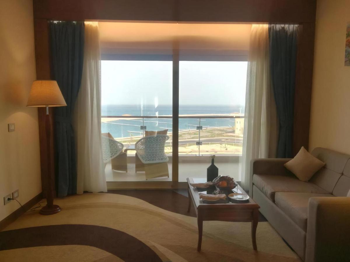 Tolip Resort El Galala Majestic Ain Sukhna Dış mekan fotoğraf The photo shows a stylish interior of a room with a large window overlooking the sea. The window features sheer and thick curtains and is framed by a balcony with two chairs visible outside. Inside, there is a comfortable-looking sofa, a coffee table
