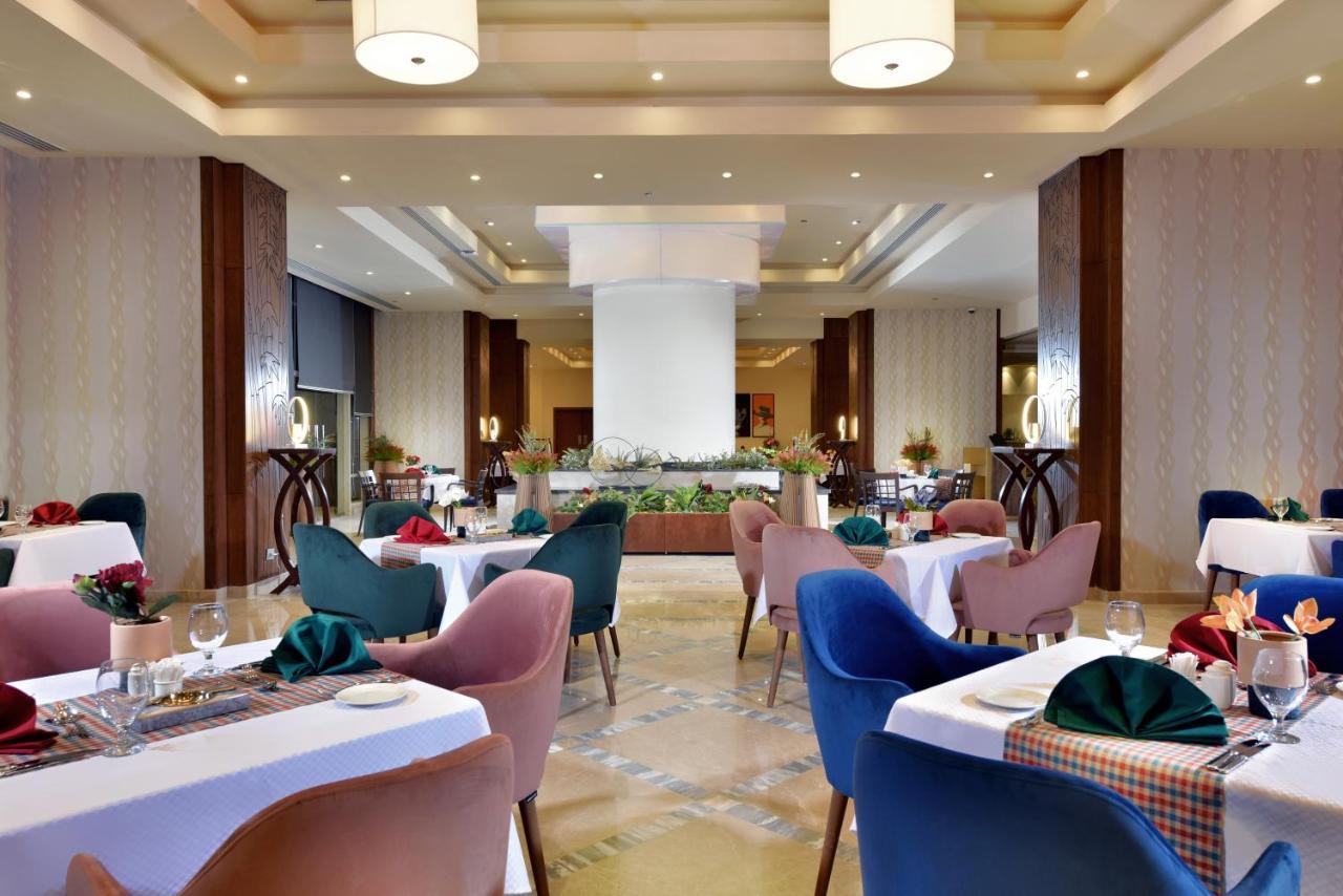 Tolip Resort El Galala Majestic Ain Sukhna Dış mekan fotoğraf The photo shows a spacious and elegantly designed restaurant interior. The layout includes several round tables dressed in white tablecloths, each set with decorative plates, cutlery, and colorful napkins. The chairs are upholstered in a variety of c