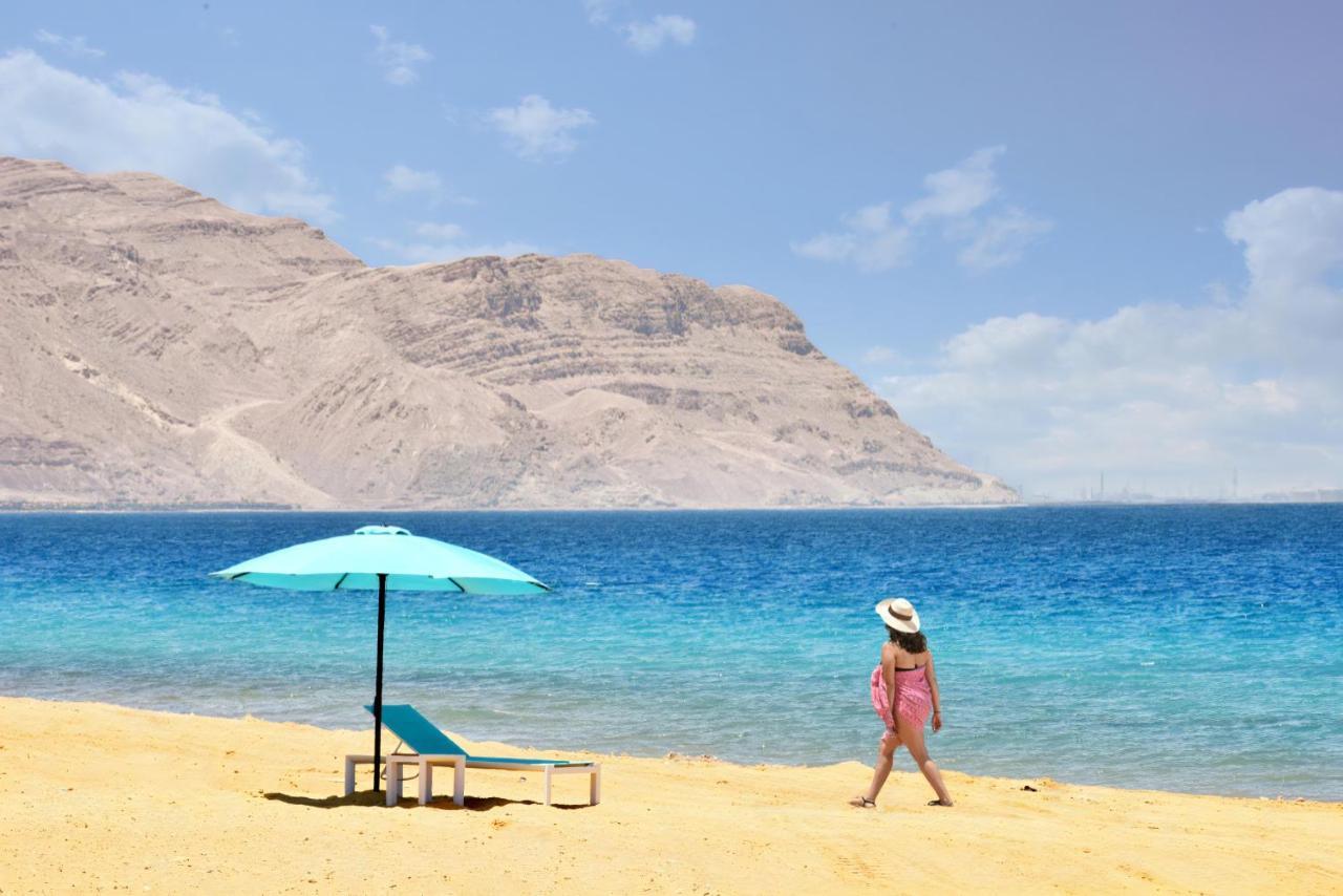 Tolip Resort El Galala Majestic Ain Sukhna Dış mekan fotoğraf The photo depicts a serene beach scene. In the foreground, there is a sandy shore with a single beach chair and an umbrella in a light blue color. A person dressed in a pink outfit is walking along the shoreline, enjoying the beach atmosphere. In the
